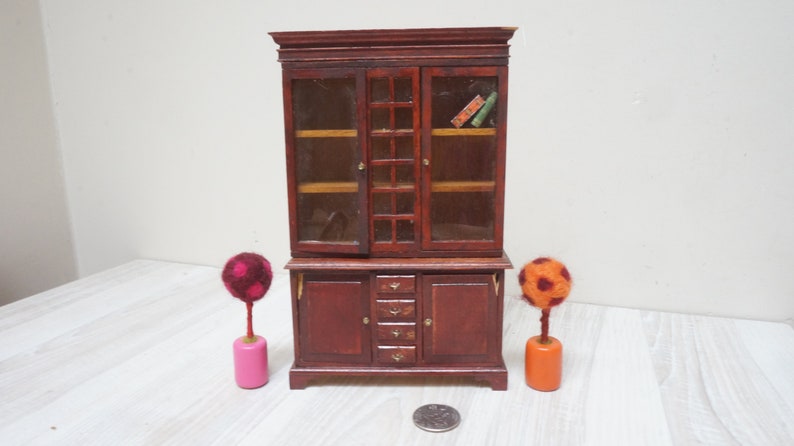 Może przedstawiać: Miniaturowa drewniana szafka z przeszklonymi drzwiami i szufladami. Szafka jest ciemnobrązowa z złotymi akcentami. Po obu stronach szafki stoją dwa małe, kolorowe drzewka z filcu.