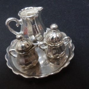 May include: A silver-colored creamer, two lidded sugar bowls, and a serving tray. The set has a decorative, scalloped edge and ornate handles. The items are made of a shiny metal.