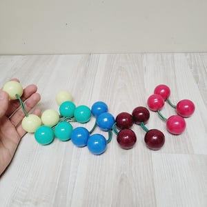 May include: A collection of colorful wooden ball toys in various shades of green, blue, red, and yellow. The balls are strung together with green cords. The toys are arranged on a light wood surface.