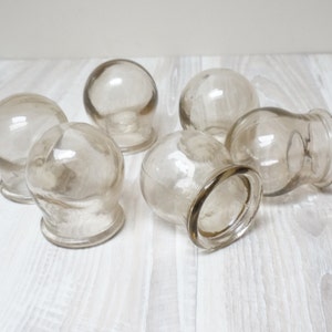 May include: Six clear glass cupping therapy cups, also known as suction cups, on a white wooden surface.