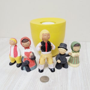 Op de afbeelding: Vijf keramische beeldjes van mensen in traditionele kleding. De beeldjes staan op een licht houten oppervlak voor een gele keramische pot.