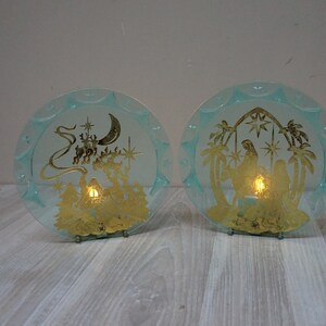 May include: Two round, light blue decorative glass plates with gold-colored religious scenes. One plate depicts a procession with a crescent moon and stars, the other shows a nativity scene. Each plate has a small light inside.