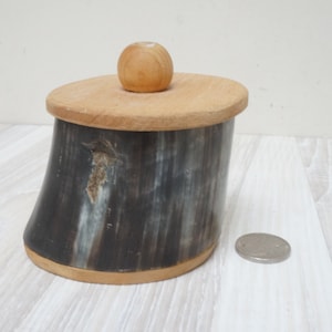Może przedstawiać: Małe, okrągłe drewniane pudełko z pokrywką. Pudełko wykonane jest z ciemnego drewna z jasnym drewnianym wierzchem i okrągłym gałką. Pudełko stoi na białym drewnianym podłożu.