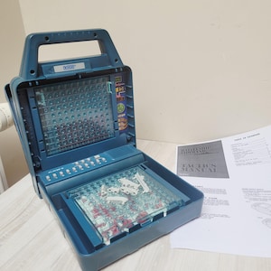 May include: A blue VTech Battleship Command electronic board game with a folding handle, a vertical grid display, and a horizontal playing surface containing white plastic ship pieces. A printed Tactics Manual document is placed to the right.