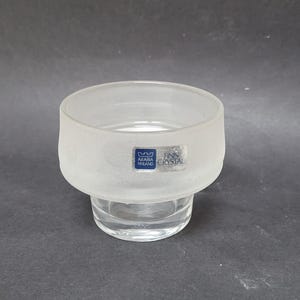 Puede incluir: Un portavelas de vidrio esmerilado con una base de vidrio transparente. El portavelas tiene el texto "FINN CRYSTAL" y un logotipo de Arabia Finlandia.