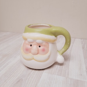 Könnte beinhalten: Eine weiße Keramiktasse in Form von Santa Clauses Kopf mit grünem Rand und Henkel. Die Tasse hat einen weißen Bart, rote Wangen und schwarze Augen.