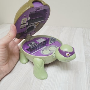 Peut inclure: Un jouet Donatello des Tortues Ninja en forme de tortue. Le jouet est vert et violet avec une coquille articulée qui s'ouvre pour révéler un intérieur détaillé. La tortue a un masque violet et est posée sur une surface en bois clair.