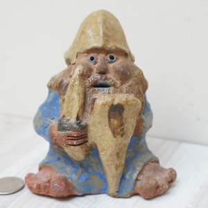 May include: A ceramic figurine of a gnome wearing a blue robe and a pointed hat, holding a sword and shield. The gnome has a white beard and blue eyes.