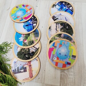 Puede incluir: Un conjunto de posavasos redondos de madera con varias imágenes. Algunos posavasos presentan paisajes urbanos, mientras que otros tienen un personaje de dibujos animados de una gota de agua con un fondo colorido. Los posavasos están dispuestos sobre una superficie de madera clara.