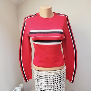 Puede incluir: Jersey rojo a rayas con rayas negras y blancas en las mangas y en el pecho. El jersey tiene una textura acanalada.