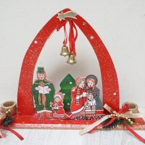 K&ouml;nnte beinhalten: Roter Weihnachtskerzenhalter aus Holz mit Glitzer-Finish. Der Halter verf&uuml;gt &uuml;ber einen festlichen Bogen mit einem Stern und Glocken sowie eine Szene mit Weihnachtslieds&auml;ngern und einem Weihnachtsbaum.