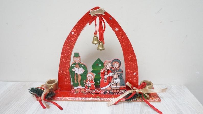K&ouml;nnte beinhalten: Roter Weihnachtskerzenhalter aus Holz mit Glitzer-Finish. Der Halter verf&uuml;gt &uuml;ber einen festlichen Bogen mit einem Stern und Glocken sowie eine Szene mit Weihnachtslieds&auml;ngern und einem Weihnachtsbaum.