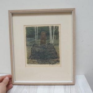 May include: A framed print of a woodblock print depicting a brown figure standing on a pile of rocks in a forest setting. The print is in a light wood frame.