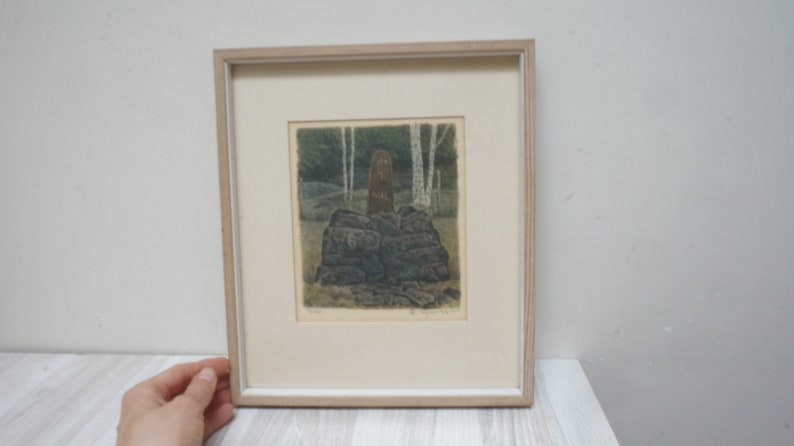 May include: A framed print of a woodblock print depicting a brown figure standing on a pile of rocks in a forest setting. The print is in a light wood frame.