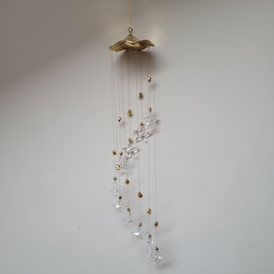May include: A decorative wind chime with a gold-colored, leaf-shaped top and hanging strands. Clear, star-shaped crystals and gold-colored beads are suspended from the strands. The wind chime is designed to hang from a ceiling or hook.