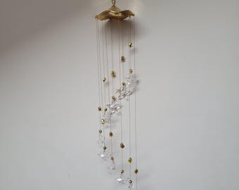Op de afbeelding: Een decoratieve windgong met een goudkleurige, bladvormige bovenkant en hangende strengen. Heldere, stervormige kristallen en goudkleurige kralen zijn aan de strengen opgehangen. De windgong is ontworpen om aan een plafond of haak te hangen.