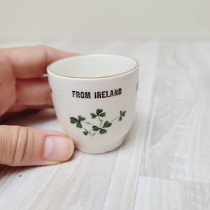 May include: A small, off-white ceramic shot glass with the text "FROM IRELAND" in black. The glass features green shamrock designs and a gold rim. The shot glass is a souvenir.