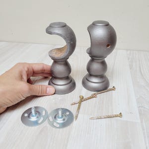Puede incluir: Dos soportes grises de madera para barra de cortina con un diseño curvo, junto con dos placas de montaje metálicas y varios tornillos dorados. Los soportes tienen un acabado metálico y una forma decorativa.