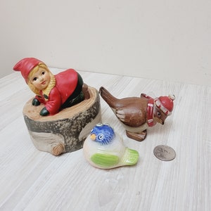 Op de afbeelding: Een verzameling keramische beeldjes, waaronder een kabouter in een rode jas en hoed die uit een boomstam komt, een bruine vogel met een rode sjaal en hoed en een kleine blauwe en groene vogel. Een munt ligt naast de vogels.