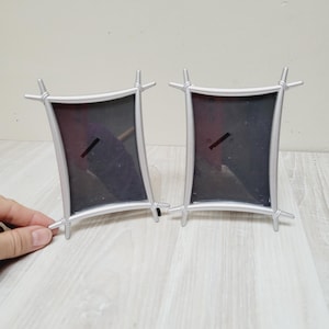 Puede incluir: Dos marcos de fotos plateados con un diseño curvo. Los marcos están vacíos y listos para mostrar fotos.