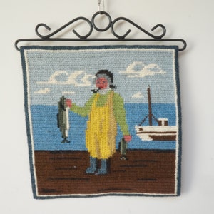 May include: A needlepoint wall hanging depicting a fisherman holding a fish in front of a boat and a blue sea with white clouds. The fisherman is wearing a yellow jumpsuit and blue boots.