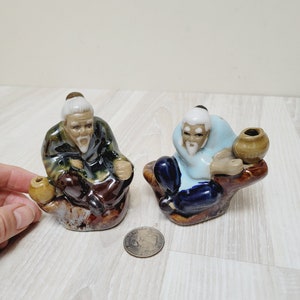 Puede incluir: Dos figuritas de cerámica de hombres mayores con barbas blancas, sentados sobre bases marrones. Una figurita lleva una túnica verde y marrón, la otra lleva una túnica azul y blanca. Ambas figuritas sostienen una pequeña olla marrón.