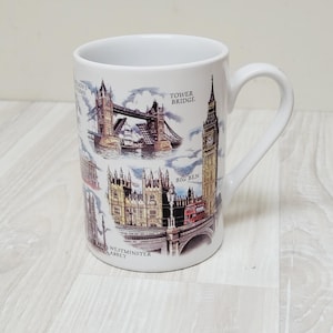 May include: White ceramic mug featuring illustrations of London landmarks, including Tower Bridge, Big Ben, and Westminster Abbey. The mug has a curved handle and is suitable for hot or cold beverages. The mug is a souvenir or gift item.