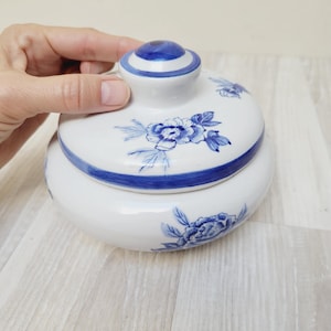 Puede incluir: Un tarro de cerámica blanco con tapa, adornado con motivos florales azules. El tarro tiene forma redondeada con una tapa con un pomo azul. El diseño floral incluye flores y hojas azules detalladas.