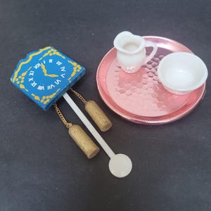 Puede incluir: Un reloj azul y amarillo en miniatura con cadenas doradas y un péndulo blanco, una jarra blanca y un tazón blanco en una bandeja rosa.