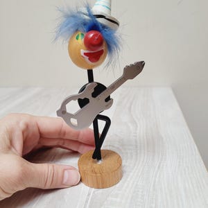 Puede incluir: Una figurita de payaso de madera con pelo azul, nariz roja y sombrero blanco y azul, tocando una guitarra plateada. El payaso está de pie sobre una base de madera.