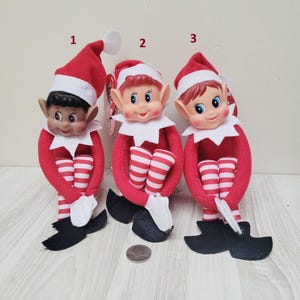 Puede incluir: Tres muñecos de elfo con piernas y brazos a rayas rojas y blancas, sombreros rojos y zapatos negros. Los muñecos tienen diferentes tonos de piel y colores de cabello. Uno tiene piel oscura, otro piel clara y el tercero cabello rojo.