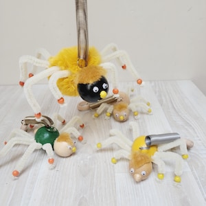 May include: A group of handmade spider toys. Each spider has a wooden body, pipe cleaner legs, and a spring. The spiders are painted in various colours, including yellow, green, and brown, with orange and yellow accents. A fun, decorative item.