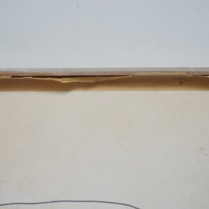 May include: A framed artwork with a white background and a thin black line drawing in the center. The frame is made of wood and has a light brown color. The frame is slightly damaged on the top edge.