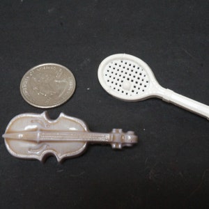 Puede incluir: Una raqueta de tenis de plástico blanco y una figurita de violín de cerámica blanca. El violín es del tamaño de una moneda de 25 centavos.