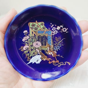 May include: A small, round, blue ceramic dish with a gold rim and a floral design featuring a cart with a roof and wheels. The cart is decorated with gold and green accents. The dish is approximately 4 inches in diameter.