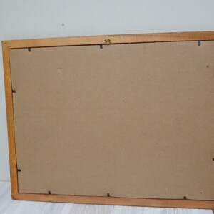 May include: A rectangular wooden picture frame with a light brown finish. The back of the frame is visible, showing a cardboard backing and small black clips to hold the contents in place. The frame is resting on a white surface.