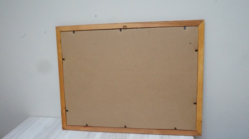 May include: A rectangular wooden picture frame with a light brown finish. The back of the frame is visible, showing a cardboard backing and small black clips to hold the contents in place. The frame is resting on a white surface.