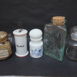 May include: Five glass and ceramic spice jars with various designs. The jars are arranged in a row on a black surface. The jars include a brown and white ceramic jar with a metal lid and the word "SPICES" embossed on the front, a white ceramic jar with a red and white lid and the word "Muskat" printed on the front, a white ceramic jar with a blue and white design, a clear glass jar with a cork lid and the word "MER" printed on the front, and a clear glass jar with a glass lid.