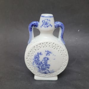 Puede incluir: Jarrón de cerámica blanca con detalles y asas azules. El jarrón presenta una escena de paisaje azul y blanco y un intrincado diseño calado alrededor del centro. Un motivo floral adorna la parte superior.