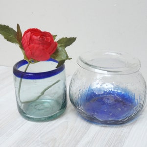 May include: Two glass vases with blue trim. The vase on the left is clear with a blue rim and holds a single red flower. The vase on the right is clear with a blue bottom and a textured surface.