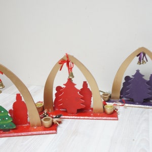 K&ouml;nnte beinhalten: Drei h&ouml;lzerne Weihnachtskerzenhalter mit B&ouml;gen, jeder mit einer Silhouette einer Familie mit einem Weihnachtsbaum. Die B&auml;ume sind rot, lila und gr&uuml;n bemalt.