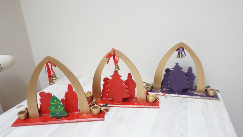 K&ouml;nnte beinhalten: Drei h&ouml;lzerne Weihnachtskerzenhalter mit B&ouml;gen, jeder mit einer Silhouette einer Familie mit einem Weihnachtsbaum. Die B&auml;ume sind rot, lila und gr&uuml;n bemalt.