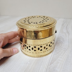 May include: A round, gold-colored metal container with a decorative pierced design. The lid features a floral-like pattern of cutouts, and the body has a series of oval cutouts. The container is held by a hand.