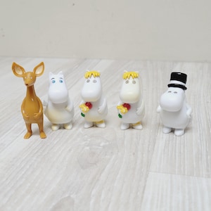 Puede incluir: Se muestran cinco figuras. Una es una criatura marrón parecida a un ciervo. Las otras cuatro son figuras blancas y redondas, dos con pelo amarillo y flores, una con sombrero de copa. Están sobre una superficie clara con vetas de madera.