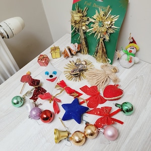 Op de afbeelding: Een verzameling kerstversieringen en decoraties. Inclusief gouden sterren van folie, rode strikken, een blauwe ster, gouden en roze ballen, een sneeuwpop en een kerstmanfiguur. Verschillende maten en vormen.