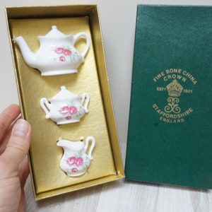 Könnte beinhalten: Ein Miniatur-Tee-Set in einer Schachtel. Das Set enthält eine Teekanne, eine Milchkännchen und eine Zuckerdose. Die Teile sind weiß mit rosa Rosenblütenmustern. Die Schachtel ist grün mit goldenen Schriftzügen, die "Fine Bone China Crown Est 1897 1901 Staffordshire England" lauten.