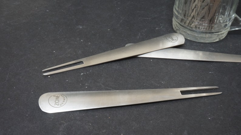K&ouml;nnte beinhalten: Zwei silberfarbene K&uuml;chenwerkzeuge aus Metall mit einem ZONE GERMANY Logo. Die Werkzeuge sind eine Gabel und ein Spatel.