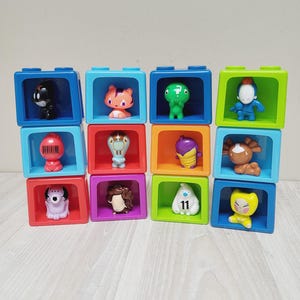 Può includere: Una collezione di dodici blocchi da costruzione in plastica colorata disposti in quattro pile verticali. Ogni blocco presenta un centro cavo contenente una piccola statuetta unica. Le figure includono varie creature, un fantasma con il numero 11 e una figura rossa con un codice a barre.