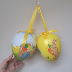 May include: Two large yellow Easter eggs hanging from a white ceiling. One egg is decorated with a chick and a basket of eggs, the other with a rabbit holding a carrot.