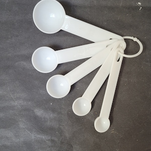 May include: A set of six white plastic measuring spoons with a ring handle. The spoons are arranged in a fan shape.